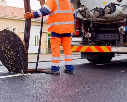 Operador com uniforme laranja e colete refletivo ao lado de caminhão de sucção em rua residencial